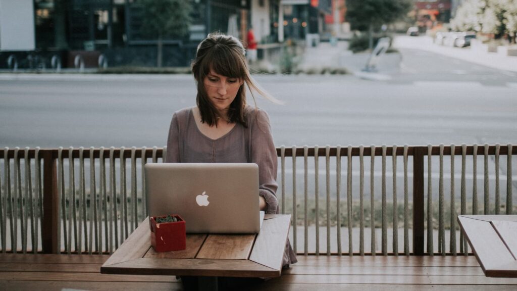 Mulher na camisa cinza sentada no banco na frente do MacBook - Curso Inglês com a Gringa com Lecil Alvino